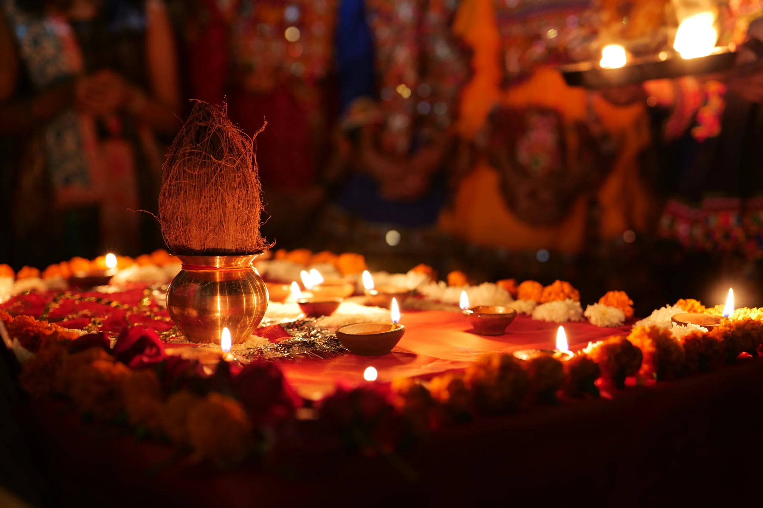 A vibrant Indian puja setup features a brass pot with a coconut, surrounded by lit oil lamps and marigold flowers, creating a warm, spiritual atmosphere.