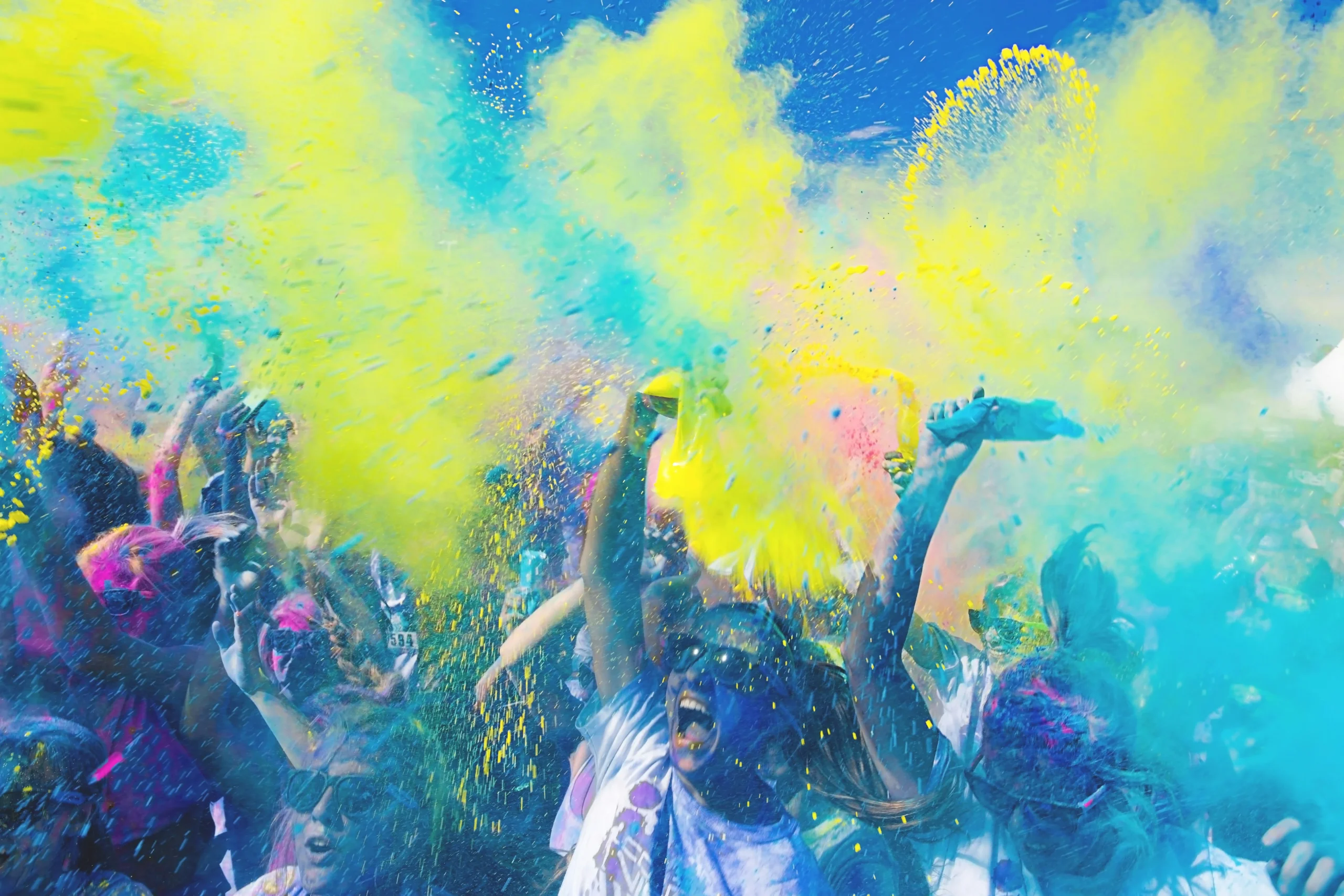 holi Colors Throwing A crowd of people outdoors celebrate Holi by throwing clouds of colorful powder, mostly yellow and blue, into the air under a bright blue sky.