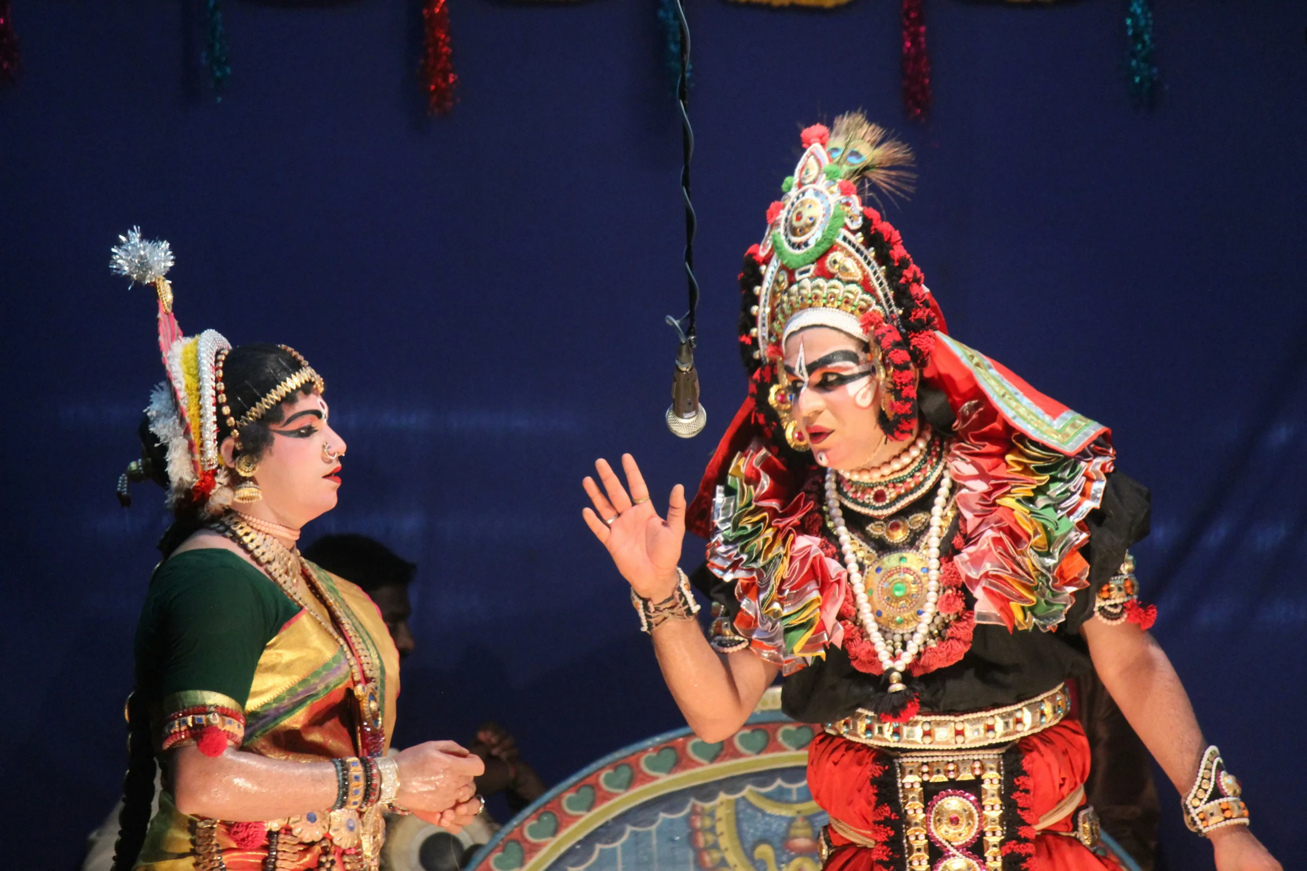 Two performers in traditional Indian Yakshagana attire and elaborate makeup, with vibrant costumes and headpieces, expressively gesturing on stage.