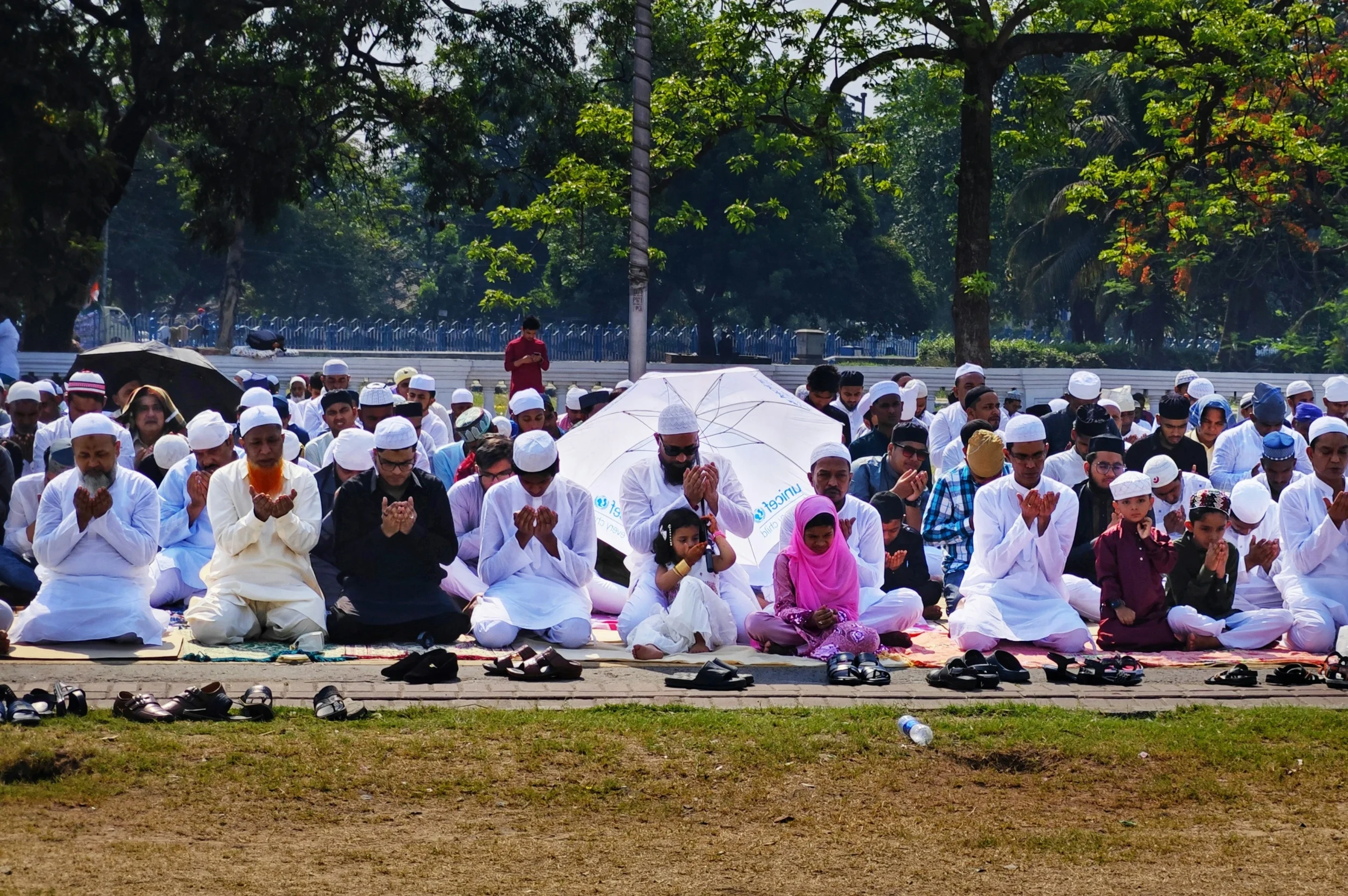 A group of people, including men and children, wearing religious attire, sit outdoors in Eid al fitr prayer. They seem peaceful, with lush green trees around.