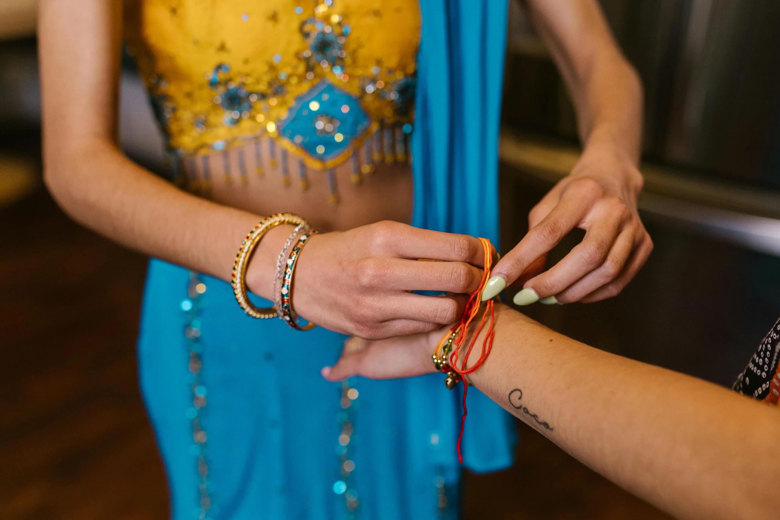 A person in a vibrant blue and yellow embellished outfit ties a red string bracelet on another's wrist. Emotion is one of care and tradition.