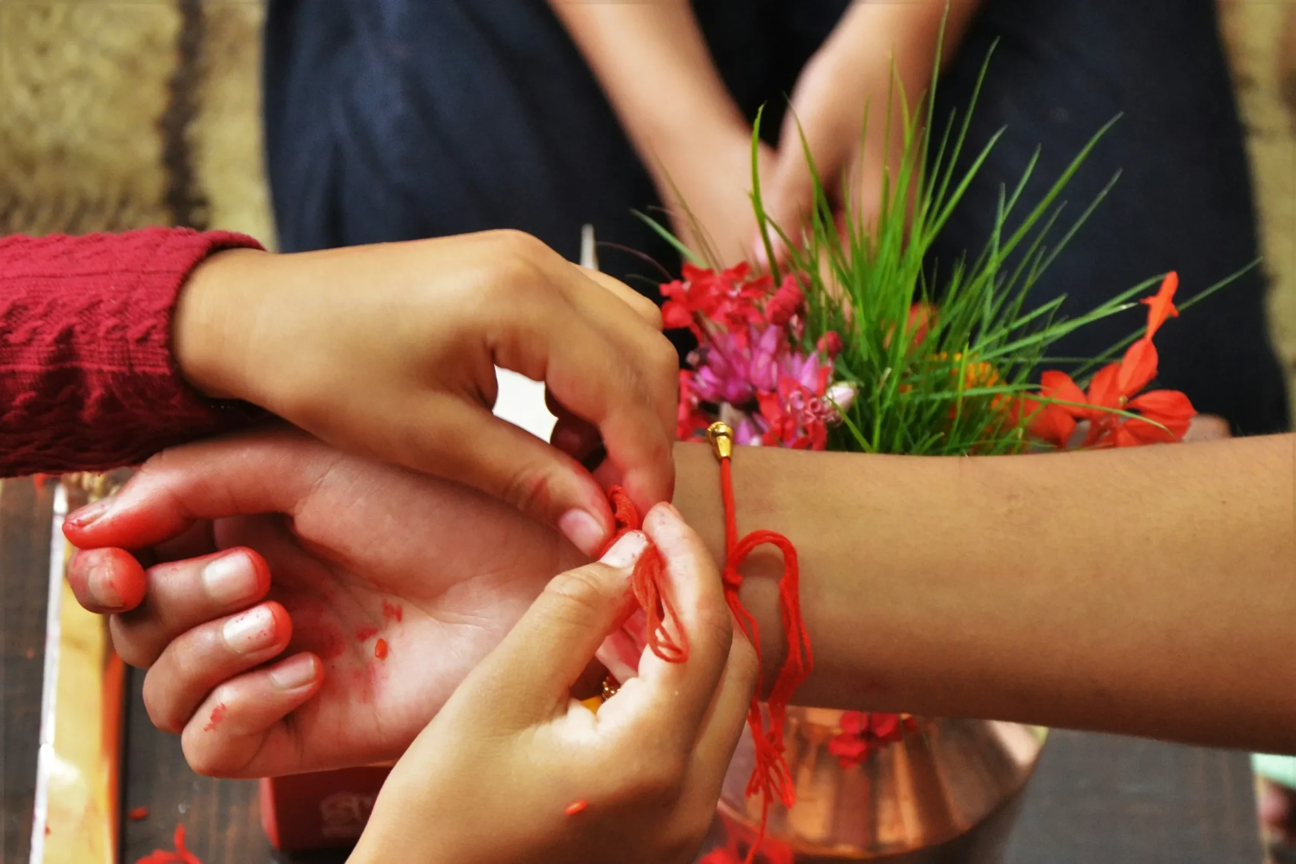 Two hands tie a red thread bracelet on a wrist, surrounded by colorful flowers and fruits, conveying tradition and ritual celebration.