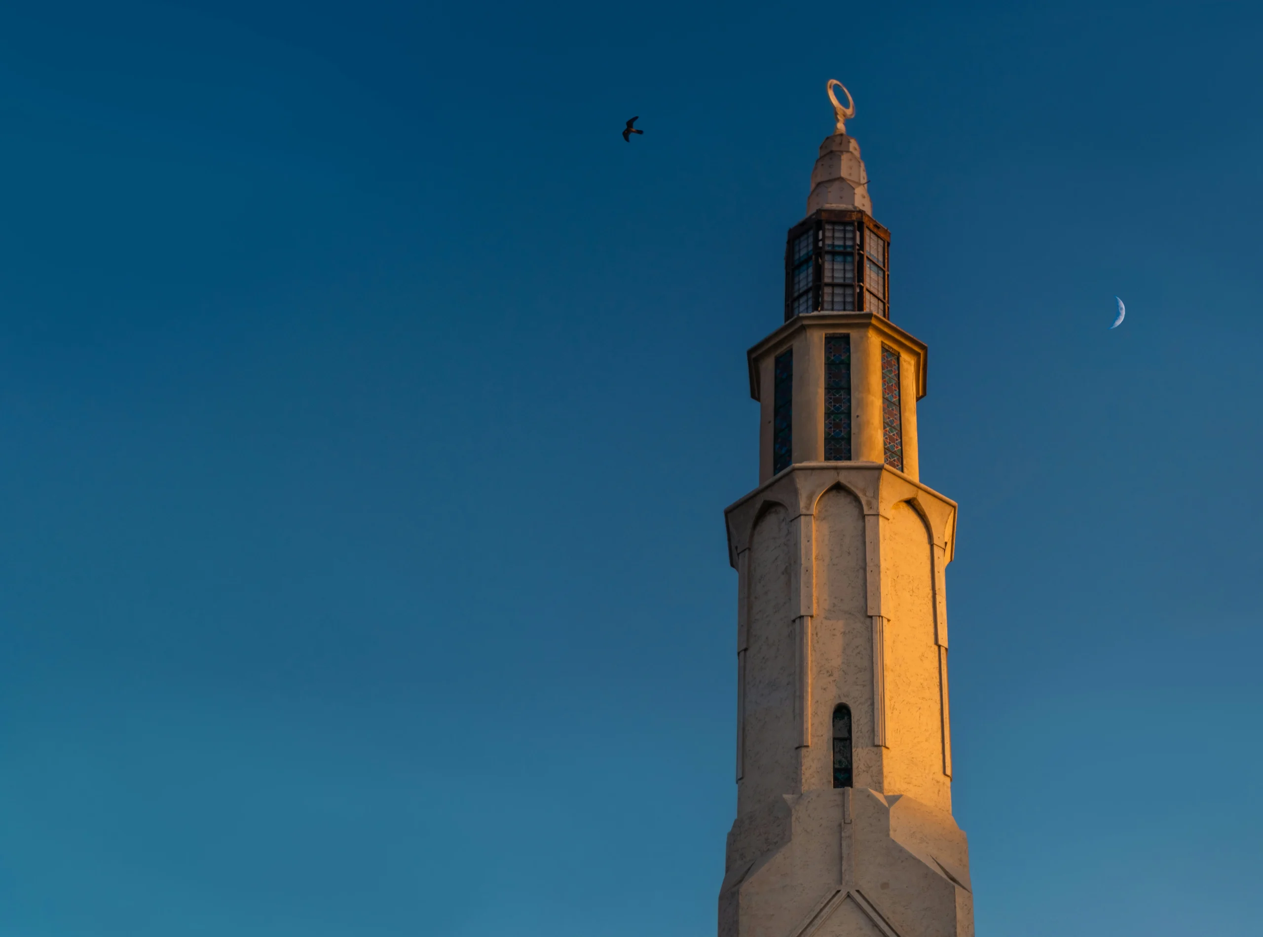 Tall minaret Tall minaret against a clear dusk sky, with a crescent moon to the right and a bird flying nearby. Warm light casts soft shadows, creating a serene atmosphere.