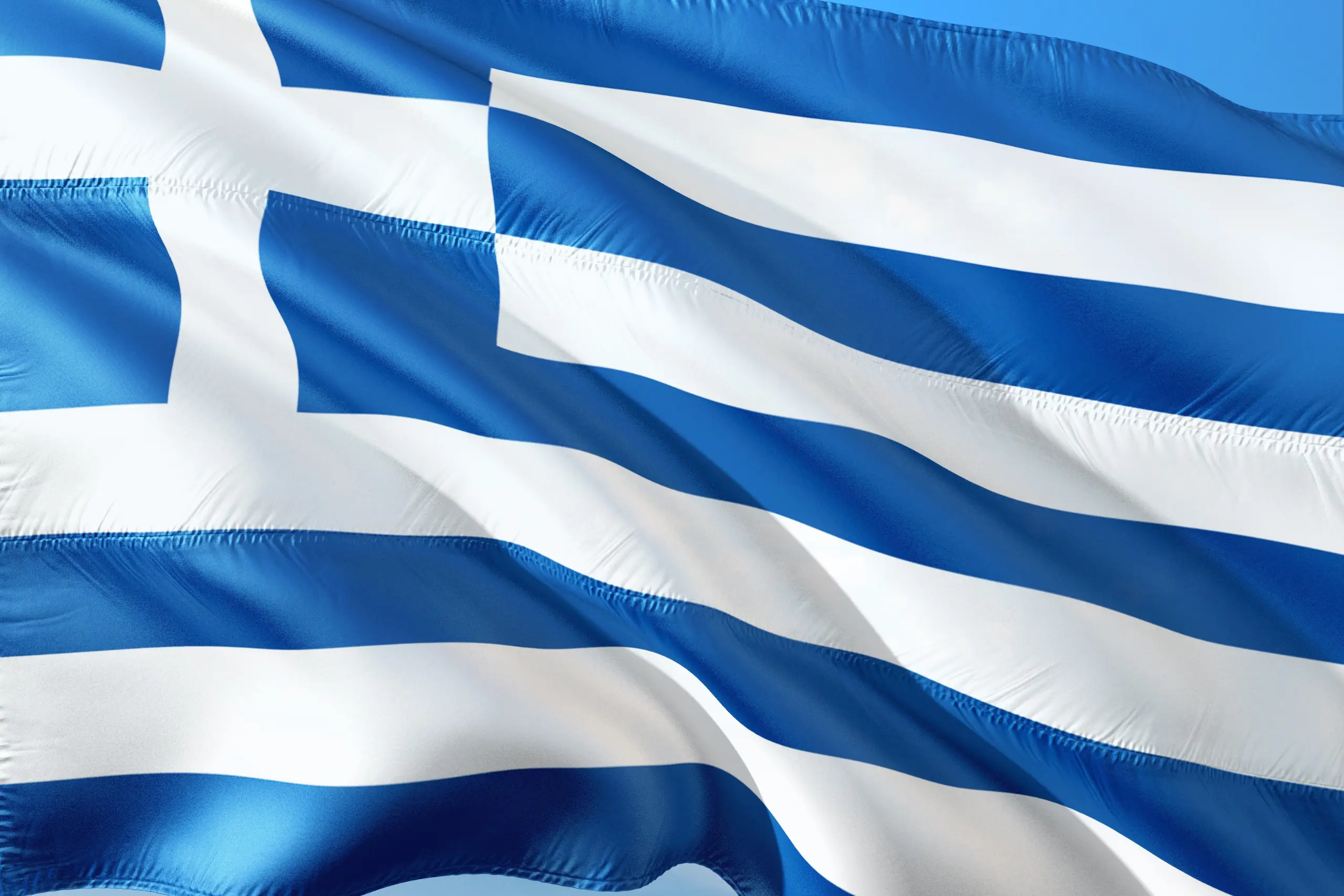 Greek national flag with blue and white stripes and a white cross on a blue square fluttering against the sky.